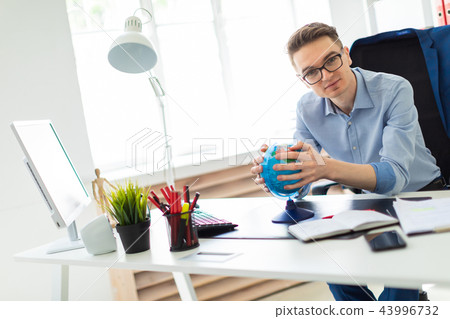 A young man sits in the office at a computer desk and holds a globe in his hands. A young man sits in the office at a computer desk and holds a globe in his hands. 43996732
