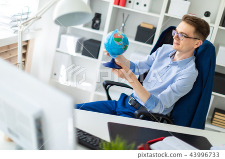 A young man sits in the office at a computer desk and holds a globe in his hands. 43996733