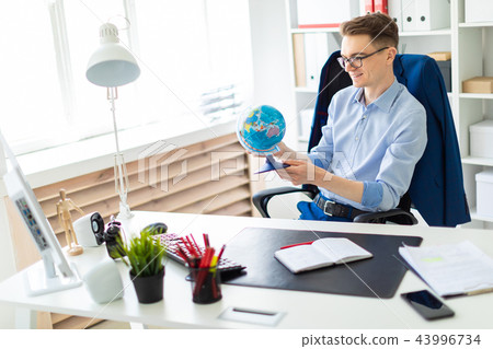 A young man sits in the office at a computer desk and holds a globe in his hands. A young man sits in the office at a computer desk and holds a globe in his hands. 43996734