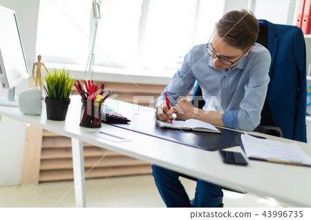 A young man sits in the office at a computer desk and writes in a notebook. A young man sits in the office at a computer desk and writes in a notebook. 43996735