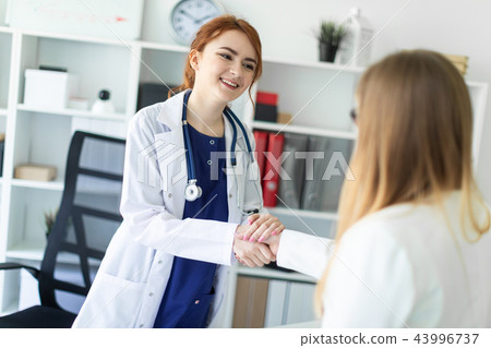A beautiful young girl in a white coat is standing near the desk in the office and shaking hands A beautiful young girl in a white coat is standing near the desk in the office and shaking hands 43996737