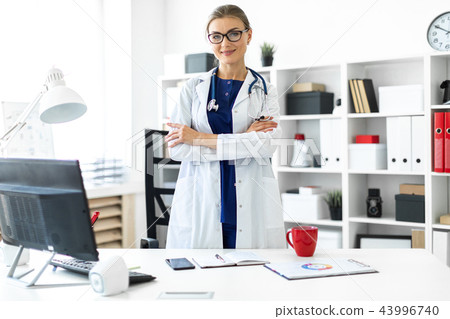 A young girl in a white coat is standing near a table in her office. A stethoscope hangs around her A young girl in a white coat is standing near a table in her office. A stethoscope hangs around her 43996740
