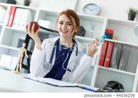 A beautiful young girl in a white robe sits at a table in the office and holds an apple and a bottle A beautiful young girl in a white robe sits at a table in the office and holds an apple and a bottle 44036259