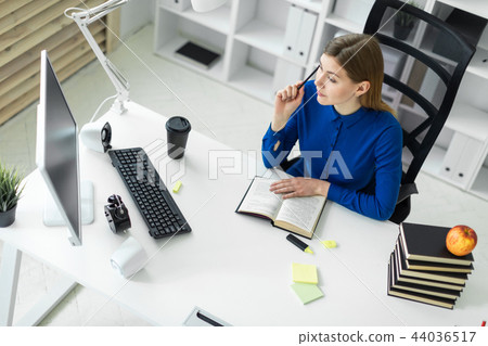 A young girl sits at a computer desk and holds a pencil in her hand. Before the girl lies an open 44036517