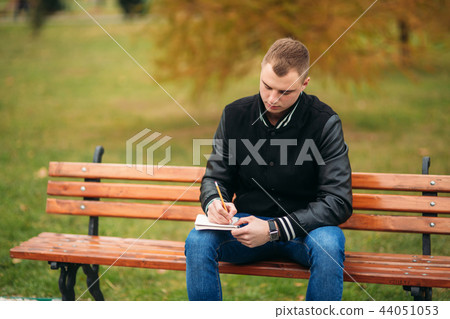 Student in a black jacket sits in a park on a bench writes down his thoughts in a notebook. Handsome Student in a black jacket sits in a park on a bench writes down his thoughts in a notebook. Handsome 44051053