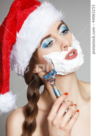 A beautiful woman in a Santa Claus hat shaves her face in a shaving foam with a man's razor. A beautiful woman in a Santa Claus hat shaves her face in a shaving foam with a man's razor. 44063255