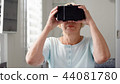 Blond senior woman in white using VR 360 glasses at home. Concept of active modern elderly people 44081780