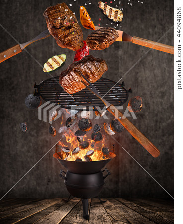Kettle grill with hot briquettes, cast iron grate and tasty beef steaks flying in the air. 44094868