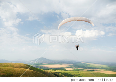 A white-orange paraglider flies over the mountainous terrain A white-orange paraglider flies over the mountainous terrain 44114058