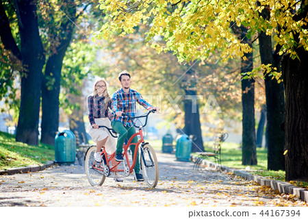 Young tourist couple, handsome man and pretty blond woman riding tandem bicycle along city street. Young tourist couple, handsome man and pretty blond woman riding tandem bicycle along city street. 44167394
