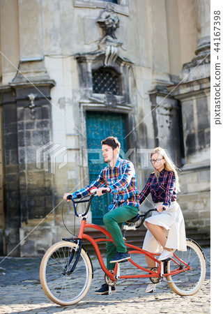 Young tourist couple, handsome man and pretty blond woman riding tandem bicycle along city street. Young tourist couple, handsome man and pretty blond woman riding tandem bicycle along city street. 44167398