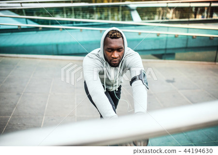 Black man runner with smartphone in an armband on the bridge in a city, stretching. 44194630