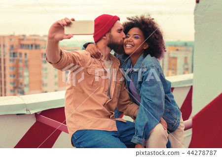 Kind man kissing his happy girlfriend while sitting on the roof and taking selfies 44278787