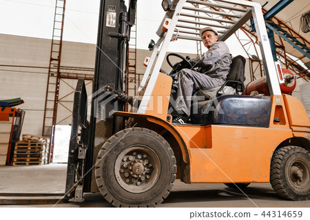 man driving a forklift through a warehouse in a factory. driver in uniform and protective helmet man driving a forklift through a warehouse in a factory. driver in uniform and protective helmet 44314659