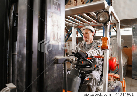 man driving a forklift through a warehouse in a factory. driver in uniform and protective helmet man driving a forklift through a warehouse in a factory. driver in uniform and protective helmet 44314661