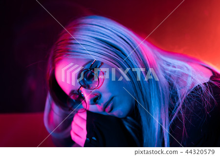 Millennial enigmatic pretty girl with unusual dyed hairstyle near glowing neon wall at night. Blue 44320579