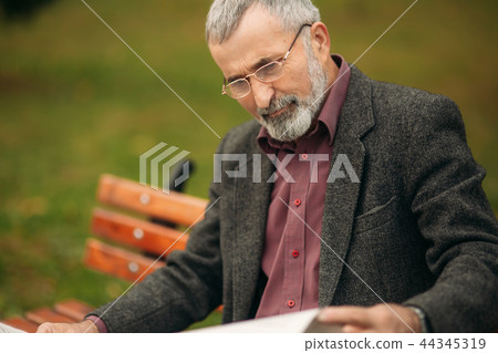 Handsome grandfather with a beautiful beard in a gray jacket sits on a bench in the park and reads a 44345319