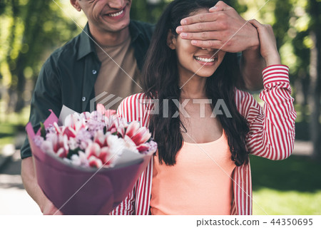 Young man closing eyes of his girlfriend and holding flowers for her Young man closing eyes of his girlfriend and holding flowers for her 44350695
