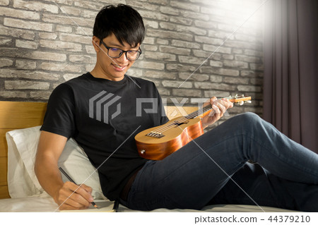 Happy young asian man playing ukulele sitting on bed in bedroom. 44379210