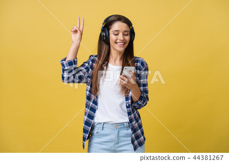 Beautiful young woman in headphones listening to music and dancing on yellow background. 44381267