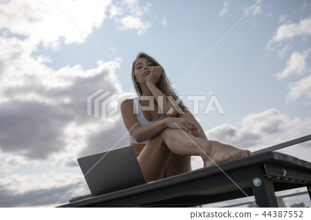 Young woman in a white top and panties sitting on the sky background and using laptop. Young woman in a white top and panties sitting on the sky background and using laptop. 44387552