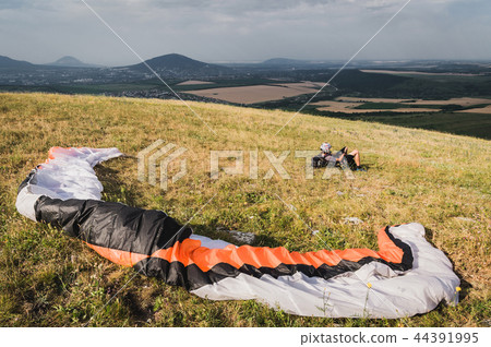 A professional paraglider in full gear and a helmet lies and rests on the grass high in the A professional paraglider in full gear and a helmet lies and rests on the grass high in the 44391995