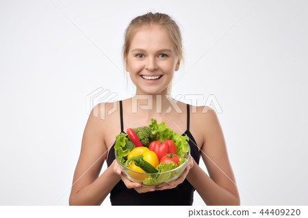 Smiling blonde young woman holding a pile of healthy fresh vegetables standing on white background. Smiling blonde young woman holding a pile of healthy fresh vegetables standing on white background. 44409220