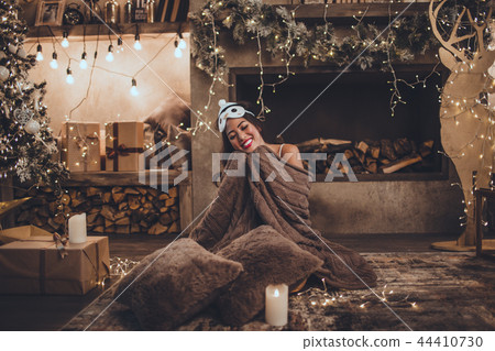 Beautiful asian woman is wearing sleeping mask and pajamas at home near christmas tree and fireplace Beautiful asian woman is wearing sleeping mask and pajamas at home near christmas tree and fireplace 44410730