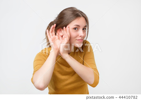 Young woman in yellow sweater listening to gossip about other with hand near ear. 44410782