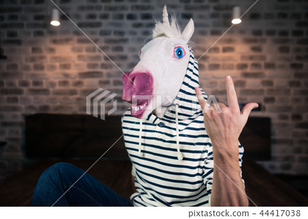 Freaky young man in comical mask sits on bed in stylish apartment. Funny unicorn Freaky young man in comical mask sits on bed in stylish apartment. Funny unicorn 44417038