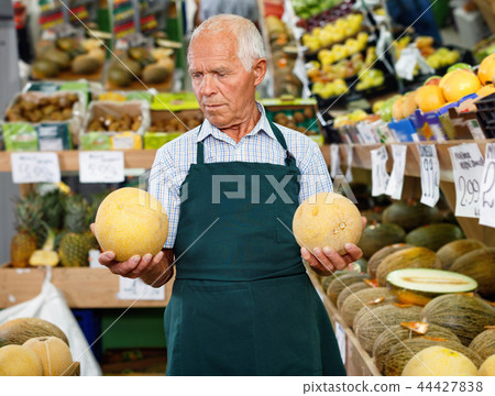 Positive senior male owner of greengrocery shop in apron offering fresh fruits and vegetables 44427838