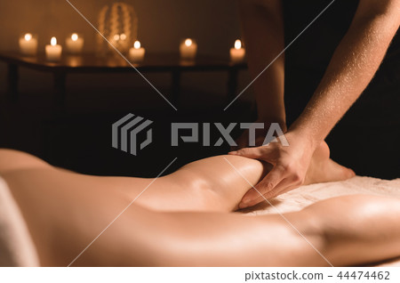 Close-up of male hands doing calf massage of female legs in a dark room with candles in the 44474462