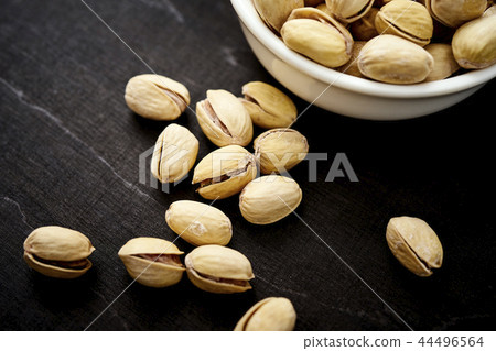 spilled delicious pistachios next to a white bowl on an old wooden black table 44496564