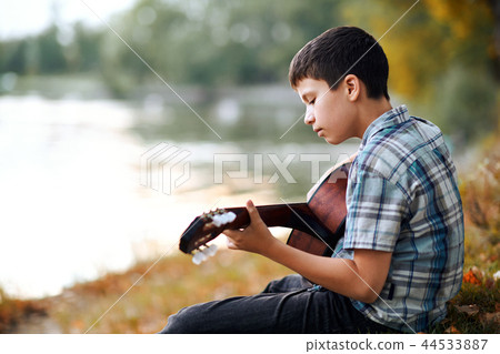 the boy plays an acoustic guitar near the river 44533887