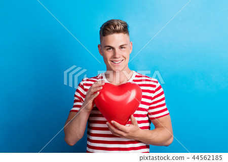 A young man in a studio, holding red heart balloon in front of him. 44562185