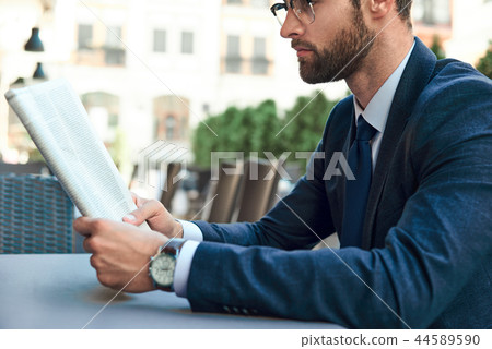 A young businessman with a beard and wearing glasses is reading a newspaper A young businessman with a beard and wearing glasses is reading a newspaper 44589590