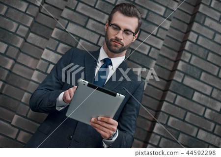 A young businessman in glasses and an expensive suit, works on a tablet in his hands A young businessman in glasses and an expensive suit, works on a tablet in his hands 44592298