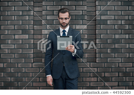 A young businessman in glasses and an expensive suit, holds a tablet in his hands A young businessman in glasses and an expensive suit, holds a tablet in his hands 44592300
