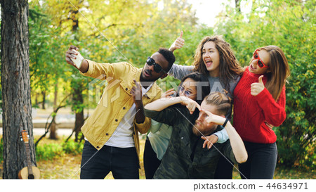 Playful youth men and women are taking selfie in park using smartphone, making funny faces and Playful youth men and women are taking selfie in park using smartphone, making funny faces and 44634971