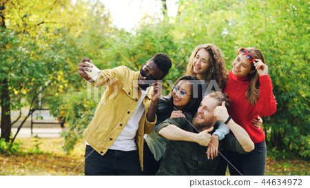 Playful youth men and women are taking selfie in park using smartphone, making funny faces and Playful youth men and women are taking selfie in park using smartphone, making funny faces and 44634972
