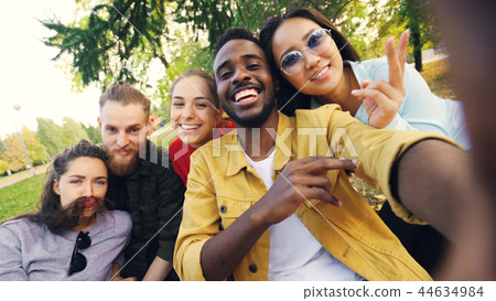 Multiracial group of friends is taking selfie in park sitting on blanket, posing and looking at Multiracial group of friends is taking selfie in park sitting on blanket, posing and looking at 44634984