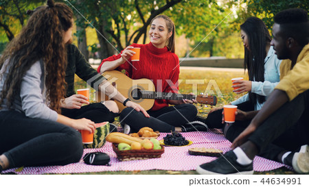 Young men and women are toasting and clinking glasses on picnic in park with guitar on warm autumn Young men and women are toasting and clinking glasses on picnic in park with guitar on warm autumn 44634991