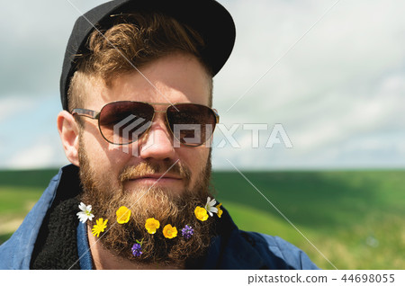 Close-up portrait of a cheerful bearded man in sunglasses and a gray cap with wildflowers in a beard Close-up portrait of a cheerful bearded man in sunglasses and a gray cap with wildflowers in a beard 44698055