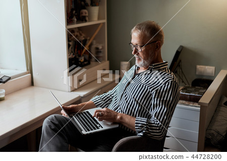 mature bearded man working from home with laptop. sitting at desk near window mature bearded man working from home with laptop. sitting at desk near window 44728200