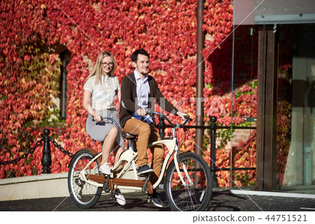 Young couple, handsome man and blond woman cycling tandem bike by building overgrown with red ivy. 44751521
