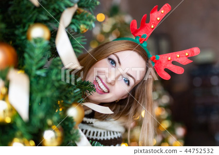 Young woman looking out of a Christmas tree wearing Christmas antlers of a deer 44752532