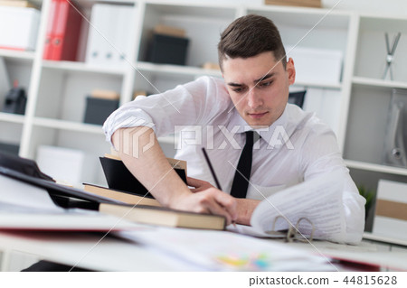 A young man working at a table in the office with a book, documents and a computer. 44815628