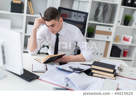 A young man working at a table in the office with a book, documents and a computer. 44815629