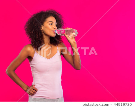 Attractive african-american sporty young woman drinking bottle of water on pink wall background Attractive african-american sporty young woman drinking bottle of water on pink wall background 44919720