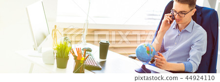 A young man sits in the office at a computer desk, talking on the phone and looking at the globe. A young man sits in the office at a computer desk, talking on the phone and looking at the globe. 44924110